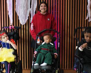 A teenager standing in a backdrop background with three younger children, who are all sitting in wheelchairs